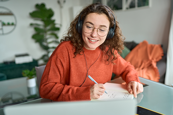 Young woman wearing headphones, taking notes while listening to an online course.